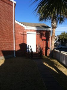 Sunny red brick building, white door, cross shadow, palm tree, St. Therese Wilmington, NC.