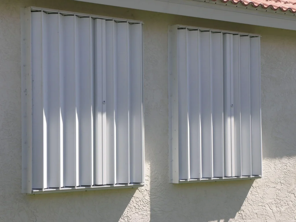 Two windows on a stucco wall are covered with TAS Approved Hurricane Shutters, offering protection from storms. Sunlight casts shadows below the shutters, highlighting their reliability for South Carolina homes. in either North or South Carolina