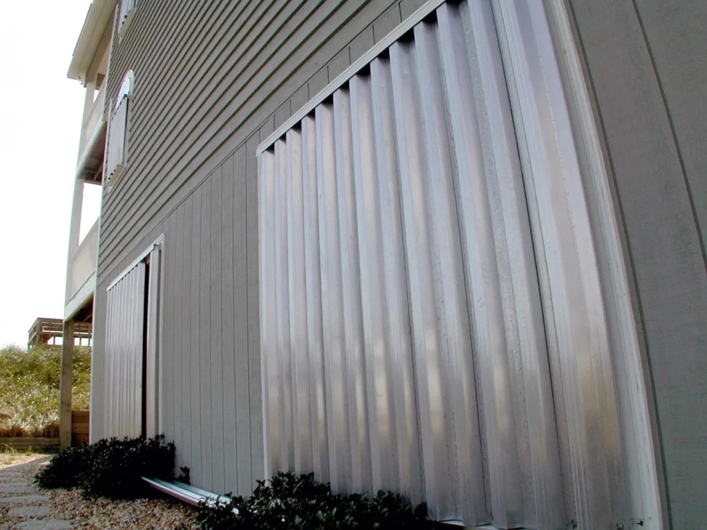 Close-up of a house with Bahama storm shutters covering several ground floor windows, providing storm protection. Small bushes line the wall below the windows. in either North or South Carolina