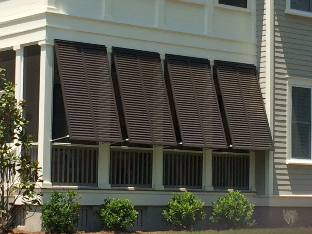 A row of five brown energy-efficient storm shutters is angled outward, shading the windows of a light-colored New Bern house with a porch and small green plants in front. in either North or South Carolina